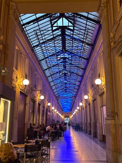 The image depicts a long, covered arcade or walkway, possibly part of a historic building in Turin, Italy. The scene is illuminated by warm, spherical lights attached to the ceiling and a vibrant blue light that emanates from the left side of the arcade, casting a striking glow on the floor and walls. On the left, a large, glass-paneled roof or window is visible, crisscrossed by a dark metal frame. Woven through this structure is an elaborate, glowing blue neon art installation that snakes and loops, creating a visually dynamic element.
Along the right side of the arcade, a row of tables and chairs is set up, suggesting an outdoor seating area for a cafe or restaurant. Several people are seated at these tables, engaged in conversation or dining. The people appear relaxed, and the atmosphere seems casual, likely indicating an evening setting due to the artificial lighting and the vibrant blue glow. The architecture of the arcade features ornate detailing on the upper walls and ceiling, with decorative molding and sculpted elements. The floor of the arcade is tiled, and the blue light creates a reflection, making the space feel expansive and modern despite the classical architecture. The time of day appears to be evening or night, as evidenced by the dominance of artificial light.