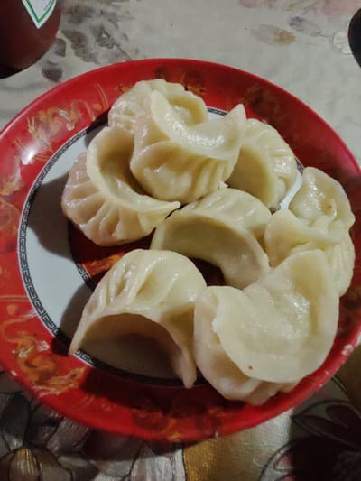 The image shows a plate of momos on a red and white patterned plate. The plate is on a table with a textured surface. In the background, a brown bottle with a green label is visible. The momos are a light yellow color and appear to be steamed. They are arranged in a circular pattern on the plate. The overall lighting is dim, suggesting the photo was taken indoors. The image is a close-up of the food, with no people visible.