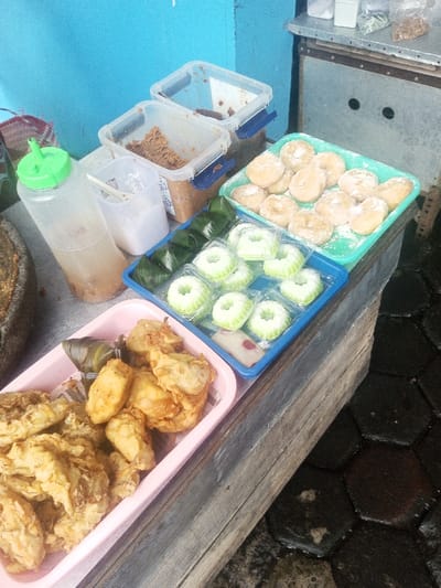Assorted Indonesian snacks are arranged on a wooden table. There are fried bananas (goreng pisang) in a pink tray. A blue tray holds green and white cakes and triangles wrapped in banana leaves. A green tray displays powdered sugar donuts. Condiments are in clear plastic containers and a squeeze bottle. The background features a blue wall and a stainless-steel cabinet. The surface below is dark, patterned tiles.