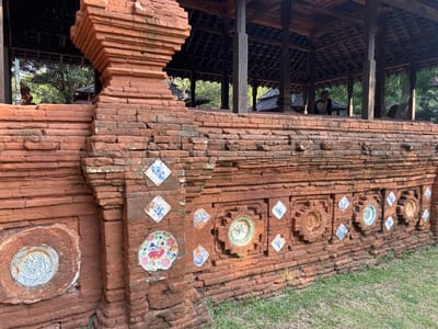 The image displays the exterior of a historical brick structure, likely part of a building or monument, in Cirebon, Indonesia. The structure is made of reddish-brown bricks and is adorned with various ceramic plates and tiles embedded within its walls. These decorations include patterned blue and white diamond-shaped tiles and larger, circular ceramic plates featuring floral and avian motifs. The architecture suggests a traditional style, with a covered wooden structure visible in the background, supported by wooden pillars. A person, appearing to be a male, is seated within this covered area, observing the surroundings. The ground in front of the brick structure is covered with green grass, indicating an outdoor setting. The overall scene conveys a sense of historical and cultural significance, typical of heritage sites.