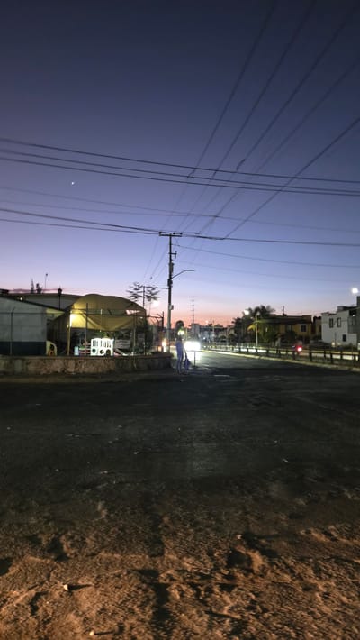 The image shows a street scene in Fraccionamiento Real del Valle, Mexico, at dusk or dawn. The sky is a gradient of purple and orange. Power lines run across the left side of the image. Streetlights illuminate a road with buildings on the left side of the road. The moon is visible in the sky. A street stand is illuminated. A person is seen near the road, standing on the right. The road takes up the right side of the image and is made of asphalt.