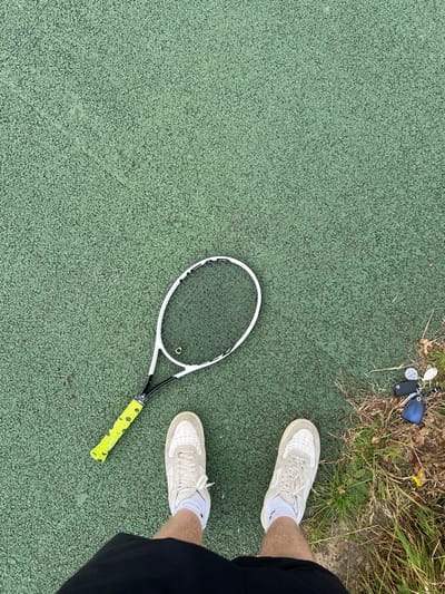 A tennis racket lies on a green tennis court. Keys rest on grass near the court's edge. A person stands on the court, wearing white sneakers and black shorts. The photo is taken from a high angle.