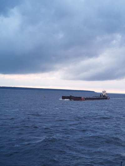 A tugboat is pushing a long, flat barge across a choppy, dark blue body of water. The sky above is overcast with heavy, gray clouds, though a sliver of lighter sky is visible on the horizon. The barge is dark red and appears to be carrying cargo covered by a gray tarp. The tugboat is white and brown and has a cabin structure at the rear. In the distance, a dark green tree line marks the shore of a landmass. A small, red buoy is visible in the water to the left of the barge. The scene appears to be set in Vichelovo, Russia.