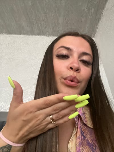 A woman with dark brown hair and long, bright green acrylic nails is looking at the camera with her lips pursed. She is wearing a light pink bracelet on her left wrist and a patterned shirt with shades of purple, gold, and black. A gold ring is visible on her left hand. The background consists of a textured white wall and a grey, textured ceiling. There is no indication of the location being Chalda, Russia.