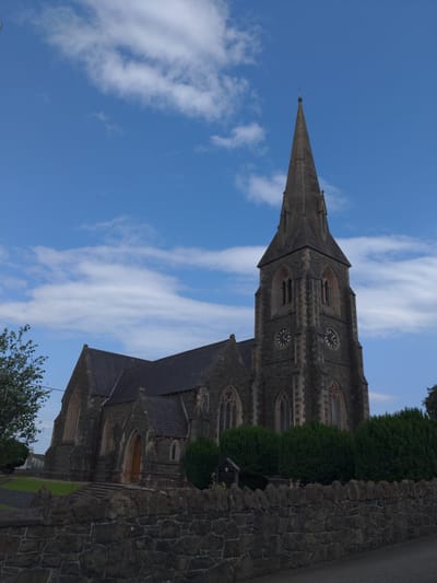 The image shows a stone church with a tall spire against a blue sky with scattered white clouds. The church has arched windows and a clock on its tower. In front of the church, there is a low stone wall and some manicured hedges. A tree is visible on the left side of the image. The scene appears to be located in the city of Magherafelt, United Kingdom. There are no people or animals visible in the image. The overall impression is one of a peaceful and well-maintained religious building.