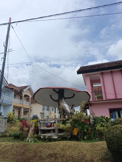 A whimsical mushroom structure stands amidst lush greenery, houses in varied pastel hues behind. The artificial tree trunk supporting a red-rimmed, white-underside mushroom cap is surrounded by flowering plants and a rustic wooden fence. A pink two-story building with a small balcony adjoins, and neighboring residences display a mix of architectural styles. Overhead, utility lines stretch across a cloudy sky.