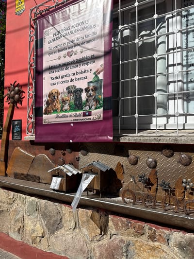 Here's a description of the image:
The image shows a section of a building exterior in Salta, Argentina. The building appears to be of mixed construction, including a window with a white security grate, a red painted wall, a section of stone masonry, and metal cladding with decorative elements.
A sign, partially obscuring the window, is in Spanish and promotes responsible pet ownership ("Huellitas de Ruhl by Cristina Gomez"), advocating for owners to pick up after their dogs. It states "Small Actions Generate Big Changes." It includes images of dogs and an illustration of a dog waste bag being deposited in a trash can.
Below the sign is what appears to be an elaborate metal sculpture or decoration. Various metal objects, including horseshoes, orbs, and what might be repurposed construction materials, are incorporated into the design. A small sign with numbers is also visible. The building is located in Salta, Argentina.