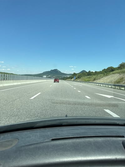 The image shows a view from inside a car traveling on a highway. The road is asphalt with white lane markings. A red car is visible in the distance, traveling in the same direction. To the left of the highway is a guardrail and a grassy embankment. To the right, there is a larger embankment covered in vegetation, with hills and mountains in the background. The sky is clear and blue. The scene is consistent with a highway in the vicinity of Zheleznovodskii, Russia, given the characteristic landscape of the Caucasus Mineral Waters region. The interior of the car, including the dashboard, is partially visible in the foreground.