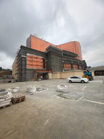 The image depicts an ongoing construction site with a large, modern building under development. The building features a distinctive copper-colored, curved facade with large windows, partially covered by scaffolding. The lower sections of the building appear to be under construction, with visible concrete and structural elements.
In the foreground and midground, the scene is dominated by a concrete surface, possibly a parking lot or work area, marked with white lines. Several large, white sacks, likely filled with gravel or construction materials, are placed on the ground. A wooden pallet with stacked materials is also visible. A white car is parked near a yellow and blue construction vehicle.
The sky is overcast with heavy clouds, suggesting a dull or potentially rainy day. The lighting is diffused and flat. The overall impression is one of active construction in a somewhat utilitarian environment. There is no visible text in the image. The setting appears to be an urban or semi-urban construction area.