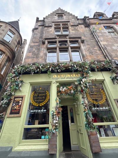 This image shows the exterior of a shop named "Arcade haggis and whisky house" located in the city of Edinburgh, United Kingdom. The shop's facade is painted a light green and is adorned with a lush garland of artificial flowers and foliage. The shop features large display windows, one of which prominently displays a circular graphic with the text "Celebrating 20 years of serving the best haggis". Another sign next to the entrance advertises "Haggis to go!". The entrance itself is a light green door, partially ajar, revealing a glimpse of the interior. Above the shop, the stone facade of an older building is visible, with traditional architectural details and several windows. Colorful triangular flags are strung across the top of the building against a cloudy sky. The overall scene suggests a festive or celebratory atmosphere for the establishment.