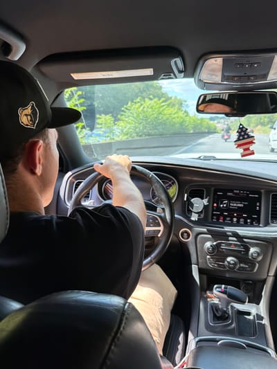 The media file captures the interior of a car from a passenger's perspective, looking towards the driver. A person, likely male, is visible wearing a black baseball cap with a logo and a black t-shirt. He appears to be the driver, with his hands on the steering wheel, though his face is mostly obscured. The scene is set within a vehicle, indicated by the dashboard, steering wheel, gear shift, and interior lights. Through the windshield, lush green trees and a bright sky suggest an outdoor setting, possibly a sunny day. A patriotic air freshener in the shape of a Christmas tree with an American flag design hangs from the rearview mirror. The car's infotainment system displays the date "10.03.2023" and the time "16:25". The overall impression is that of a casual car journey.