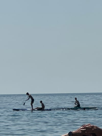 In the waters off Đurovići, Montenegro, three individuals are engaged in water activities. Two people are on paddleboards, one standing and paddling, the other seated. A third person is partially visible in the water, possibly having fallen from a board. The water is calm with gentle ripples, reflecting the clear, pale sky. The scene appears to be a peaceful daytime setting.