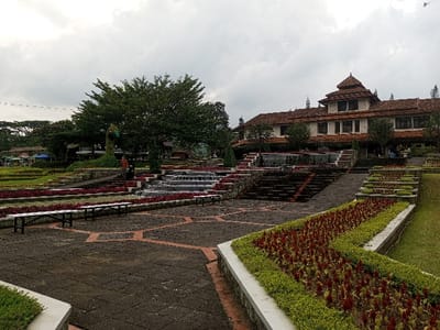 A tiered garden landscape under a cloudy sky features flowerbeds filled with red blooms alongside stepped water features. Geometric pavement leads toward benches and landscaped levels. A large tree stands near the center, and a multi-story building with a tiered roof is visible in the background. The overall scene suggests a meticulously designed park or public space.