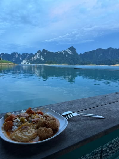 The image depicts a meal served on a white plate placed on a weathered wooden surface. The plate contains rice, what appears to be fried chicken or fish balls, sliced carrots, and a fried egg, all coated in a light orange sauce. A fork and spoon are set next to the plate.
The setting is a waterfront, with a calm, blue body of water stretching towards a mountainous landscape in the distance. The mountains are covered in dense green vegetation. The sky is a clear blue with some wispy clouds. The overall impression is that of a tranquil outdoor dining experience.
The image is oriented vertically, with the food plate and wooden table in the foreground, and the water and mountains forming the background. The lighting suggests it is daytime, likely late morning or afternoon, with bright, diffused sunlight. The location is identified as Khao Phang, Thailand, a region known for its scenic lakes and limestone karsts.
There is no visible text in the image. The scene evokes a sense of relaxation and enjoyment of a meal in a beautiful natural setting.