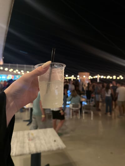 In Seaside Heights, United States of America, a person's hand with light-colored fingernails holds a clear plastic cup filled with a light-colored liquid and ice, with a black straw. The cup is covered in condensation. The background is blurred, showing a crowd of people standing and mingling under a dark ceiling with visible lighting fixtures. Several tables are scattered throughout the venue.