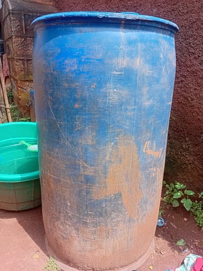 A blue plastic barrel lies horizontally on top of two green plastic basins, outdoors, near a wall in Ndeiya, Kenya. The barrel appears used and scratched, with some dirt or rust staining its surface. The surrounding environment includes a brown, earthy surface, some vegetation, and a reddish-brown textured wall.