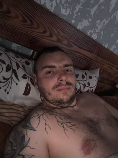 The image shows a man lying in bed, facing the camera. He has short dark hair, a beard, and a mustache. He is wearing a silver chain around his neck. Tattoos are visible on his left shoulder and chest, including a tree-like design. The man is positioned on a pillow with a brown floral pattern, against a backdrop of a wooden headboard and a textured wall. The lighting appears dim, suggesting it may be nighttime or a dimly lit room. The scene is indoors, likely in a bedroom. There is no information to indicate this scene is located in Częstochowa, Poland.