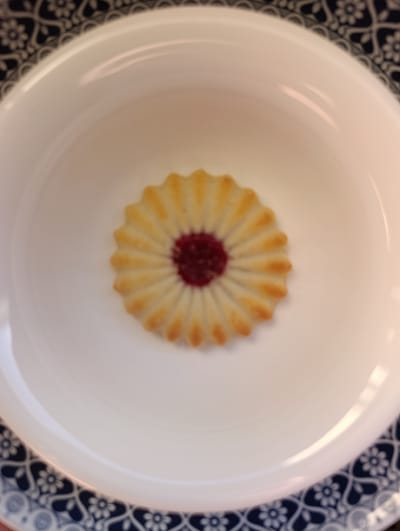 This media file shows a close-up overhead shot of a single shortbread cookie with a jam filling, placed in the center of a white bowl. The cookie has a fluted, circular shape with a reddish-pink jam in its center. The bowl is a plain white ceramic, and it rests on a plate with a decorative blue and white pattern. The pattern consists of floral motifs. The lighting suggests it might be daytime, possibly indoors, with soft light highlighting the cookie and bowl. There is no visible text in the image. The setting appears to be domestic. This image is contextualized as originating from Nablus, Palestinian Territories.
