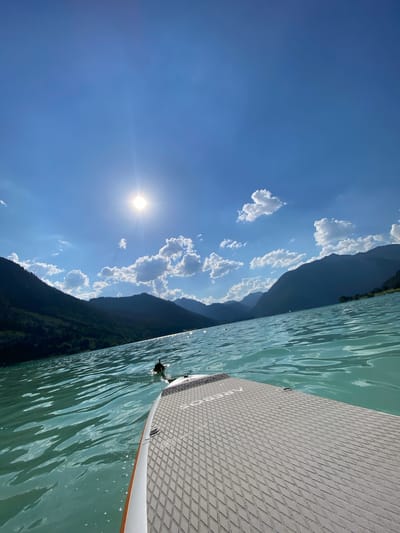 The image depicts a serene outdoor scene on a bright, sunny day. The primary subject is a stand-up paddleboard positioned on clear, turquoise water. The paddleboard's textured surface, designed for grip, is visible in the foreground, with its edge gently touching the rippling water. Beyond the paddleboard, the water stretches out, reflecting the bright sky and the surrounding landscape.

The background is dominated by a vast, deep blue sky dotted with fluffy white clouds. The sun shines brightly, casting a lens flare and illuminating the scene with a warm glow, suggesting it is midday. Along the horizon, dark, verdant mountains rise steeply from the water's edge, their slopes covered in dense forests. The combination of the clear water, the bright sun, and the majestic mountains creates a peaceful and picturesque atmosphere.

The image evokes a sense of tranquility and enjoyment of nature. The low angle perspective, likely taken from someone on the paddleboard, immerses the viewer in the experience. The lack of visible people suggests a moment of solitary enjoyment or a quiet outing. The overall impression is one of a beautiful, leisurely day spent on the water in a stunning natural setting. The visible text "RED PADDLE" is faintly visible on the paddleboard. The context provided mentions Lärchenwiese, Austria, hinting at a location in the Austrian Alps, known for its pristine lakes and mountainous terrain.