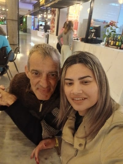 A selfie-style photograph captures a woman and a man posing closely together. The woman has light brown hair with blonde highlights, a fair complexion, and is wearing a light-colored jacket. The man, positioned behind her, has gray hair and appears older. The background reveals an indoor setting resembling a restaurant or cafeteria. Tables, display cases with food items, and other people are visible, suggesting a public gathering place. The photo is somewhat blurry, indicating possible low light conditions or motion during capture.