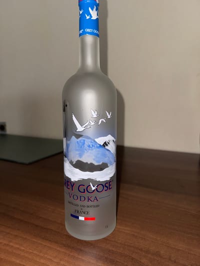 A single 1-liter Grey Goose vodka bottle stands upright on a polished, medium-brown wooden surface, likely a table or desk. The bottle is made of frosted glass, featuring a blue foil seal on its neck with white goose motifs and "GREY GOOSE" branding. Its body displays an illustration of white geese flying over blue and white mountainous terrain. Below the illustration, the "GREY GOOSE" logo is prominently displayed in red and blue, followed by "VODKA," "DISTILLED AND BOTTLED IN FRANCE," a stylized French flag, and the "1L" volume indicator.

In the background, a plain, light-colored wall, possibly beige or off-white, extends across the frame. To the left of the bottle, a dark, flat, rectangular object, possibly a desk mat or glass panel, is visible on the wooden surface, partially obscured by the bottle. A dark, indistinct object rests upon this flat surface further in the background. The lighting is uniform, suggesting an indoor setting. There are no individuals, overt actions, or interactions depicted in the image. The scene is located in Kumanovo, North Macedonia, although specific establishment details or exact location within the city are not visually discernible.