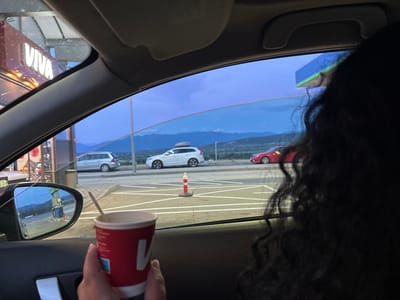 The image shows a person holding a red and white disposable coffee cup with a wooden stir stick, inside a vehicle. The person's hand and forearm are visible on the right side of the cup. The vehicle's interior is dark, with part of the dashboard and a door handle visible. Through the car window, a gas station is visible with a sign displaying "VIVA" in white lettering. Multiple cars are parked at the gas station and on the road beyond. The sky is a deep twilight blue, with mountains visible in the distance. The scene appears to be at a gas station located in Kalugerovo, Bulgaria.