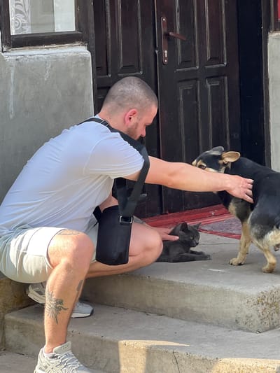A man is outdoors, interacting with a dog and a cat near a dark wooden door. He is wearing a white t-shirt, light-colored shorts, and athletic shoes. A black bag is slung across his shoulder. The dog, a mix of black and tan, is standing and being petted by the man, with its head turned towards him. A grey cat is lying down on a concrete step, with one of the man's hands visible near it. The scene appears to be in daylight, likely late morning or early afternoon, given the bright sunlight casting shadows. The setting includes a concrete stairway and the wooden door of a building. The man's tattoo is visible on his lower leg.