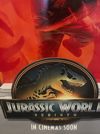This media file is a photograph of a movie poster for "Jurassic World: Rebirth." The poster features a stylized, metallic emblem with a dinosaur skull. To the left of the emblem, a person's legs and arm are visible, dressed in clothing that appears to be a light grey jumpsuit. The background on the left is a vibrant orange gradient, with shadows suggesting movement or a dynamic scene.
The emblem is circular with a golden border. Inside, a golden silhouette of a dinosaur head is prominently displayed against a dark, textured background. The teeth are sharp and the eye socket is clearly defined.
To the right of the emblem, the movie title "JURASSIC WORLD" is displayed vertically in large, metallic silver letters with a dark outline. Below the title, the word "REBIRTH" is smaller and stacked. Further to the right, the text "IN CINEMAS SOON" is visible in white, capital letters. The overall aesthetic is dramatic and conveys a sense of anticipation for the film's release. The lighting appears to be studio-like, highlighting the textures and metallic finishes of the poster elements. The image seems to be taken indoors, likely in a cinema lobby or a promotional space. There are no clear indicators of time of day, weather, or specific emotions, beyond the implied excitement of a movie premiere. The location context of Byala Slatina, Bulgaria is noted but not visually evident within the poster itself.