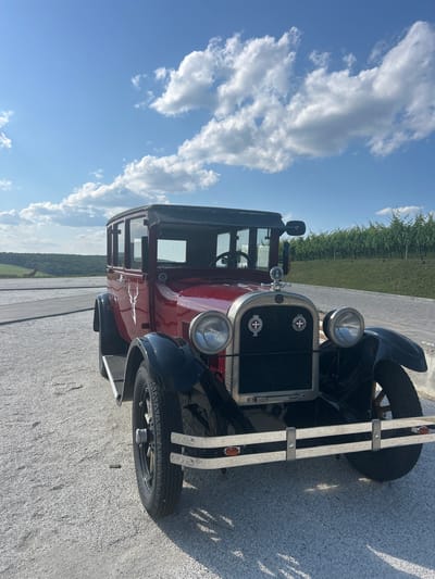 A vintage red automobile with black fenders and roof is parked on a gravel lot. The car has a chrome grille and bumper, two round headlights, and a spare tire on the side. A white deer silhouette emblem is visible on the driver's side door. In the background, a vineyard stretches across a grassy hillside under a bright blue sky with scattered white clouds. To the left of the car, a wooden boardwalk leads towards a distant, tree-lined landscape with a few brown horses grazing. The scene appears to be located in the vicinity of Ulmu, Moldova.