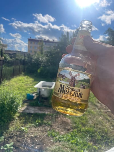 A person's hand holds a glass bottle of "Старый Мельник" (Old Miller) beer. The label features a vintage illustration of a windmill and a horse-drawn cart, with text that includes "из БОЧОНКА" (from the barrel). The bottle contains a golden-colored liquid. The background suggests an outdoor setting on a sunny day. The sky is blue with fluffy white clouds, and sunlight is visible, creating lens flare. Green grass and trees are in the background, along with a building and what appears to be a white tub and a blue bin. The overall scene suggests a casual moment outdoors, possibly in a backyard or rural area. The time of day appears to be late morning or afternoon due to the bright sunlight. The weather is clear and sunny.