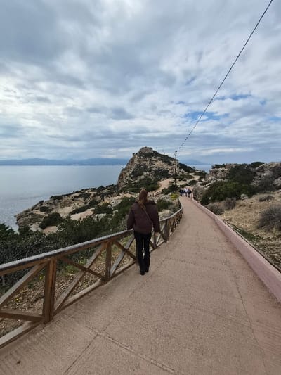 The image displays a paved coastal pathway under an overcast sky in the Municipal Unit of Loutraki - Perachora, Greece. A single individual, viewed from behind, with long light brown hair, wearing a dark brown jacket, black trousers, and carrying a dark shoulder bag, walks along the path. To the left, a wooden railing with horizontal and diagonal members separates the path from a steep, rocky slope covered in sparse green and brown vegetation that descends to a calm, dark blue-grey body of water. A prominent rocky promontory, topped with indistinct structures, extends into the water in the mid-ground, with hazy mountains visible on the distant horizon. Further along the pathway, near the promontory, a small group of approximately three to five individuals is also walking away from the camera. The light brown, textured path is bordered on its right side in the mid-distance by a low, light pink concrete barrier. A dark, thin utility line stretches diagonally from the upper right of the frame towards the center-left. The sky is dominated by layers of grey and white clouds.