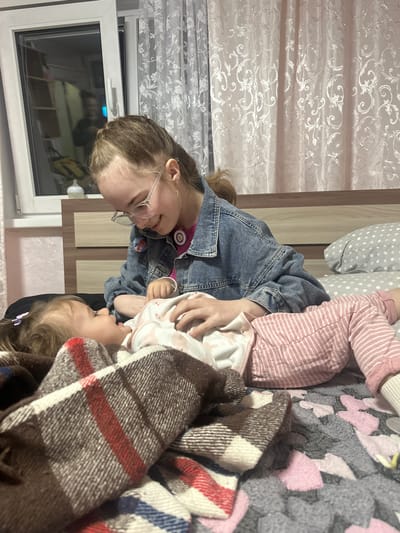 Two Caucasian females are interacting on a bed in an indoor setting. The older female, wearing glasses and a denim jacket, is facing and gazing at a younger female who is partially covered by a brown, white, and red plaid blanket. The younger female is lying down. The bed has a patterned grey and pink blanket, and the person above the older female is wearing pink and white striped pants and is lying on a grey pillow with white dots. A window with white blinds is visible. The location is Uva, Russia.
