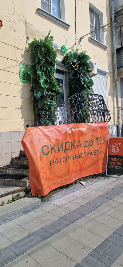 The image shows a storefront in Miass, Russia, featuring a large orange banner that reads "СКИДКА до 15% НА ГОТОВЫЕ БУКЕТЫ" (Discount up to 15% on ready-made bouquets). The banner is draped across a black metal railing. The storefront is decorated with lush green artificial plants. To the left of the entrance, a green sign displays a tulip logo and the hours "8:00 - 22:00". To the right of the entrance, a smaller dark green sign with a tulip logo reads "СВЕЖИЕ ЦВЕТЫ ОТ fanfan tulipan" (Fresh flowers from fanfan tulipan). The building has a light yellow facade with some visible cracks. Steps lead up to the entrance, and the ground is paved with grey and darker grey tiles. The scene appears to be a retail establishment, likely a flower shop, on a sunny day.