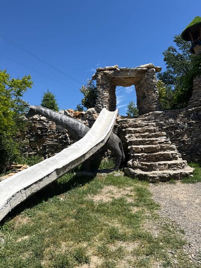 The image shows a stone structure in Lukovit, Bulgaria. A concrete slide is in the foreground, leading down from the stone structure. A stone archway is visible above the slide. The environment includes green grass, trees, and a blue sky.