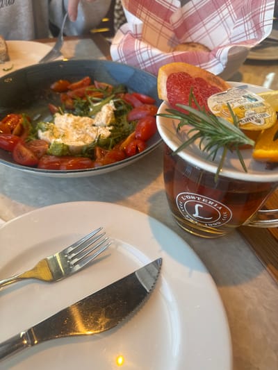 This image depicts a table setting within a restaurant identified as L'Osteria, located in Mysiadło, Poland. The scene is primarily focused on food and beverages. In the immediate foreground, a white ceramic plate holds a silver fork and a silver serrated knife, positioned horizontally. To the left, a dark blue bowl contains a caprese-style salad consisting of halved red cherry tomatoes, fresh green arugula, and several portions of white soft cheese, possibly burrata or mozzarella, with traces of green pesto. To the right, a clear glass mug, branded "L'OSTERIA L EST. 1999", holds an amber-colored liquid, likely tea. Resting on top of the mug, on a small white dish, are garnishes including a wedge of pink grapefruit, a sprig of fresh rosemary, a small plastic honey container labeled "GRETSAMER HONIG RIVERGOLD", a lemon wedge, and an orange wedge. In the blurred background, a hand wearing a gray sleeve holds a fork near another plate, suggesting the presence of at least one other individual. Further back, a bread basket lined with a red and white checkered napkin contains a single bread roll. The table surface is a light-colored, possibly marble, material.