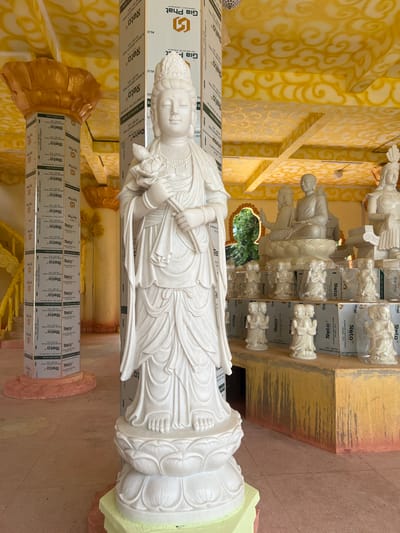 An indoor religious facility in Đăk Mar, Vietnam, is depicted, dominated by a tall, white marble statue of a female deity, likely Guanyin, standing centrally and facing forward. The figure, adorned with an elaborate headdress and flowing robes, holds a lotus flower in her left hand and stems in her right, resting on a multi-tiered lotus pedestal. The ceiling and upper architectural elements are painted yellow and gold with swirling cloud patterns. Support pillars in the foreground and mid-ground are covered with white aluminum composite panels featuring "Gia Phat Alu" and "Reto" branding. To the right of the central statue, a raised platform with a discolored yellow-and-brown finish supports multiple white statues. These consist of larger seated Buddhist figures on lotus bases and multiple smaller statues, some enclosed in clear plastic, depicting praying or gesturing figures arranged in rows. An arched opening in the distant background provides a view of green foliage. The floor consists of light-colored, patterned tiles. To the far left, a golden pillar with a lotus-shaped capital and yellow decorative elements are present.