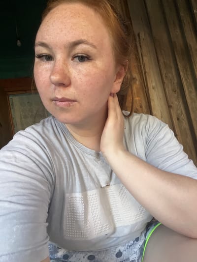 A young woman with fair skin and red hair is shown in a close-up portrait. She has freckles scattered across her face, prominent eyelashes, and her hair is pulled back. She is wearing a light gray t-shirt with a textured pattern on it and what appears to be patterned shorts with a neon green trim. Her right hand is touching her neck. The background consists of weathered wooden planks, suggesting an indoor or rustic setting. No specific location in Аряж, Russia is identifiable from the image.