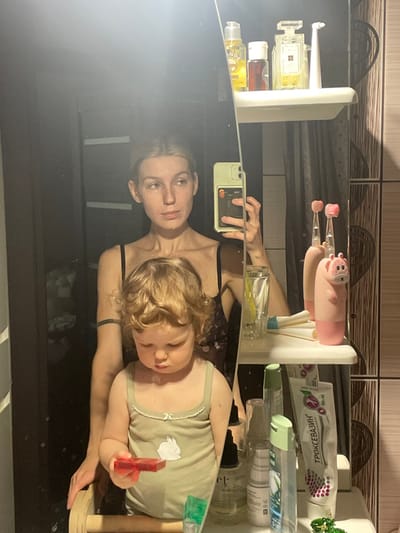 The image shows a reflection of a woman and a young child in a bathroom mirror. The woman is holding a smartphone in her right hand and looking into the mirror. She is wearing a black tank top with floral print and a black tattoo on her left arm. The child is holding a red toy and looking down. The child is wearing a green tank top with a white bunny on it. There are shelves on the wall with numerous toiletry items on them. This bathroom is located in Moscow, Russia, at the coordinates 55.659 latitude and 37.591 longitude.