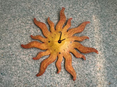 A sun-shaped clock is mounted on a textured, light blue wall. The clock has a yellow face with orange and red rays extending outwards. The numbers 3, 6, 9, and 12 are visible on the clock face. The hour hand points to the 9, and the minute hand points to the 12. The second hand is orange and points slightly past the 9. The clock appears to be a decorative item. The background is a mottled blue surface with a rough texture, possibly wallpaper or a painted finish. There is no indication of the city of Osentsy, Russia in the image.