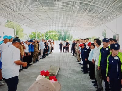 The image depicts a solemn gathering of people, likely in a covered outdoor area with a corrugated metal roof supported by white beams. A long, narrow stretcher with two handles is placed on the ground, adorned with red and white flowers. Two lines of individuals, predominantly young men and some women, stand facing each other, forming an aisle. Most are wearing uniforms or casual attire, including hats and caps. On the left side, a person in a white shirt and dark pants is prominent in the foreground. On the right side, individuals wear dark blue uniforms with fluorescent green accents, and some wear red caps. The background shows greenery and trees, suggesting an outdoor setting that is shaded or covered. The presence of the stretcher and flowers indicates a ceremony or memorial event. The scene is set in Karagana, Uzbekistan, although no specific landmarks identify the exact location within the city.
