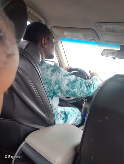 A man is seen from behind and the side, driving a car. He has short, dark hair and a beard, and is wearing a green and white patterned shirt. He is holding the steering wheel with both hands. The car's interior is visible, including black leather seats, a center console, and a rear-view mirror. A sun visor with a warning label is also visible. The image is taken from inside the car, looking forward. There is a watermark "Q Express" in the bottom left corner. The setting is implied to be the city of Sabojibi, Nigeria.
