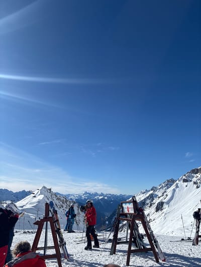 The image depicts a snow-covered alpine environment on a clear, sunny day, located within the general vicinity of Klösterle, Austria. The sky is deep blue, with bright sun glare visible in the upper left quadrant. The foreground features a flat, snow-covered area where several individuals in ski attire are present. Two wooden ski racks, holding multiple pairs of skis and poles, stand in the center and right foreground. One rack displays a red and white sign with "WINTERSPORT" partially visible.
In the foreground, a person in a black jacket and red pants, wearing a white helmet, stands with their back to the camera, beside a ski rack. Another person, largely obscured, in a red jacket is seated or leaning at the bottom center. In the midground, a person in a red jacket and dark helmet stands holding ski poles near a rack, facing slightly right. To their left, another individual in a light blue jacket and helmet stands with skis and poles. Additional figures in ski gear are partially visible further back.
The background consists of extensive snow-covered mountain ranges under a clear sky. A prominent, jagged, snow-capped peak is visible in the mid-left, with a ski lift structure or station at its summit and an associated lift line extending down. A valley containing a distant settlement is discernible in the mid-right. The slopes on the right side of the frame feature steep, rocky terrain covered in snow, with visible avalanche control fencing. Blue flags or markers are scattered across the snow in the midground.