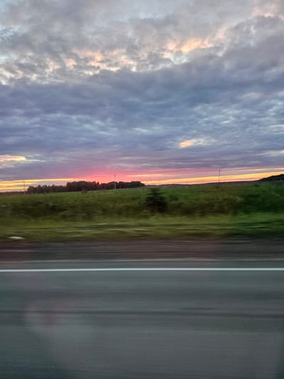 The image shows a sunset over a rural landscape. The sky is filled with dramatic clouds, tinged with pink and orange from the setting sun. Below the sky, a vibrant band of pink and orange stretches across the horizon, marking the sun's position. A dark line of trees forms a silhouette against the colorful sky, with a green field extending in front of them. The foreground of the image is dominated by a blurred asphalt road with a white line marking a lane. The motion blur suggests the photo was taken from a moving vehicle. The scene appears to be located outside of Большая Уса, Russia, given the rural setting. There are no people or animals visible in the image.