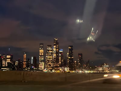 The image depicts the Manhattan skyline at night, viewed from Weehawken, New Jersey, United States of America, looking eastward across the Hudson River. The sky above the cityscape is dark and partially obscured by clouds. Numerous skyscrapers and buildings form the distant skyline, heavily illuminated with a predominant array of warm yellow and orange lights from windows and exterior features, complemented by scattered cool white, blue, red, and green lights. In the mid-ground, a substantial, indistinct, light brown concrete barrier or embankment spans horizontally across the lower third of the frame. In the immediate foreground, blurred streaks of light from moving vehicles are visible, specifically an orange/yellow light trail and a white light trail on the lower right, indicating active roadway traffic. Lens flares or reflections, appearing as vertical streaks of light, are present in the upper right quadrant of the image. The entire scene is characterized by soft focus. No individual people or explicit actions are discernible.
