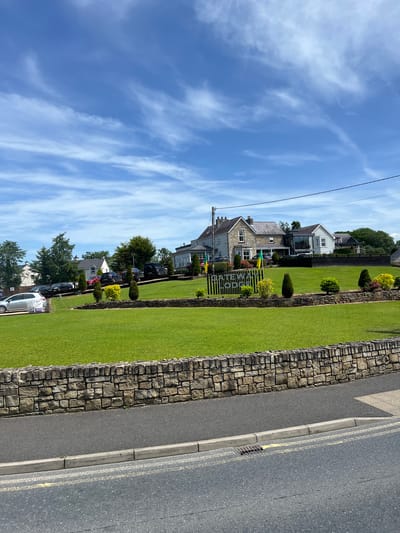 This is a medium-shot, vertically oriented photograph of a street in Donegal, Ireland. The scene depicts a row of houses with well-maintained lawns, bordered by a stone wall and a paved road. A bright blue sky with wispy white clouds dominates the upper portion of the image.

The houses appear to be residential, with a mix of architectural styles. The foreground features a neatly mown green lawn and a decorative stone wall. Beyond the wall, a paved road with yellow double lines is visible, suggesting it is a public thoroughfare. Cars are parked along the side of the road, indicating a residential area. In the middle ground, a sign with the text "GATEWAY LODGE" is clearly visible.

The time of day appears to be midday or early afternoon, given the bright sunlight and clear sky. The weather is pleasant and sunny. The presence of a stone wall and the style of the houses suggest a location in Ireland, consistent with the provided location context. There are no people visible in the image, and the overall mood is one of tranquility and order. The image captures a typical street scene in a residential area on a fair day.
