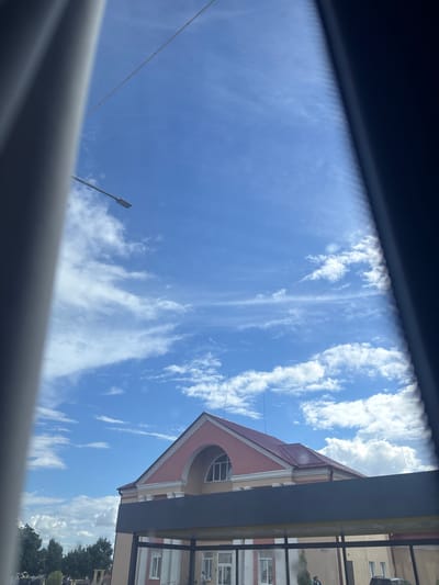 This image captures a view of a building with a pink roof under a bright blue sky with scattered white clouds, possibly taken from within a vehicle. The building appears to have arched windows and decorative columns. In the foreground, dark frames or structures partially obscure the view. The environment suggests an outdoor setting in a town or city, with trees visible in the distance. The scene is illuminated by daylight. The provided metadata indicates the location is in Капыльскі сельскі Савет, Belarus.