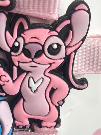 A close-up, eye-level shot shows a pink cartoon character, possibly a charm or toy, with large eyes, ears, and a tail. The character has a black outline and is made of a rubbery material. It is attached to a pink woven ribbon. The background is white. The image does not provide any information about its location in the city of Ravno pole, Bulgaria, or any details about people, actions, or interactions.