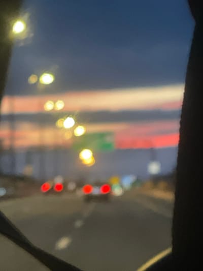 This is a blurry, out-of-focus photograph taken from inside a vehicle, likely at dusk or night. The sky displays hues of deep blue, with streaks of pink and white, possibly indicating a sunset or twilight. Several bright, circular lights are visible, suggesting streetlights or headlights. In the foreground, the taillights of other vehicles appear as red circles. A blurry green sign, possibly a road sign, is visible in the distance. The overall impression is one of movement and urban ambiance, captured at low light conditions. The photograph was taken in Pesochny, Russia.