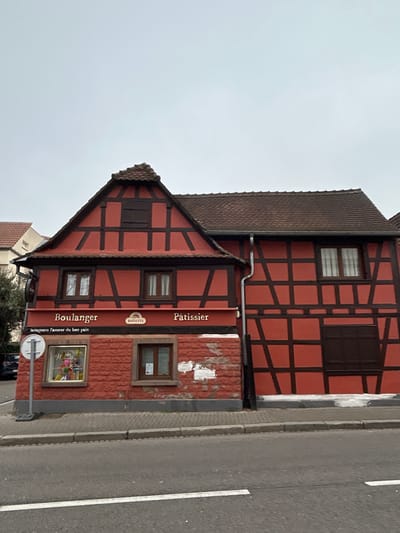 The image displays a two-story, half-timbered building situated in Hoenheim, France, under an overcast sky. The structure features a dominant red exterior with dark brown timber framing and dark brown tiled roofs. The left section of the building presents a gabled roofline with a small, dark-shuttered attic window. Below this, two white-framed windows are visible on the first floor. A prominent red horizontal sign on the ground floor identifies the establishment as a "Boulanger" (bakery) on the left and "Pâtissier" (pastry shop) on the right, with a "BANETTE" logo centrally placed. Below "Boulanger," the text "Artisans l'amour du bon pain" is present. The ground floor facade consists of reddish-brown textured masonry with several patches of white, possibly peeling render or paint. A large display window on the left exhibits various colorful items, while a smaller, brown-framed window is to its right. The right section of the building maintains the red and dark brown half-timbered style, featuring one visible window on the first floor and a set of dark brown shutters covering an opening below. A silver-colored downspout runs vertically on the right side of the left building section. In the foreground, a grey asphalt road with a single white solid lane marker runs horizontally, bordered by a grey concrete sidewalk and curb. A circular white traffic sign with a black horizontal line is positioned on the sidewalk to the left. No individuals are visible, and no actions or interactions are depicted. To the far left, a portion of another building with a light-colored facade and some dark green foliage is partially visible.
