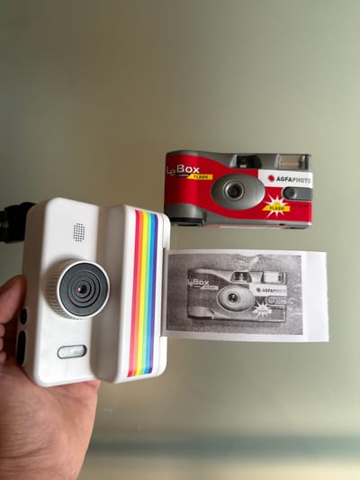 This image shows two cameras and a printed photograph. In the foreground, a white instant camera with a rainbow stripe design is held by a person's hand. The camera has a prominent lens, a small speaker grill, and a flash button. To the left and slightly above, a red and silver "AGFAPHOTO Le Box Camera Flash" is visible. A black and white photograph is being printed from the white instant camera, showing an image of the red and silver camera. The background is a neutral, reflective surface. The scene appears to be indoors, possibly in a home or studio, with soft lighting. No specific time of day or weather conditions can be determined. The text visible includes "AGFAPHOTO", "Le Box CAMERA", and "FLASH" on the red camera, and a timestamp "2023/07/23 09:38:17" on the printed photograph. The overall impression is one of reviewing or demonstrating the functionality of the cameras.